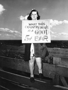 Photo of a girl sitting on a fence, holding a sign that says "What This Country Needs is a Good 5 Cent Bar"