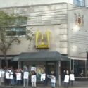 A photograph of workers with picket signs standing outside the Bay Building downtown, with the A-Channel "A" logo above them.